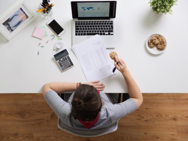 woman with tax report eating cookie at office business, accounting and freelance concept - woman with calculator, papers and laptop computer eating cookie at office table